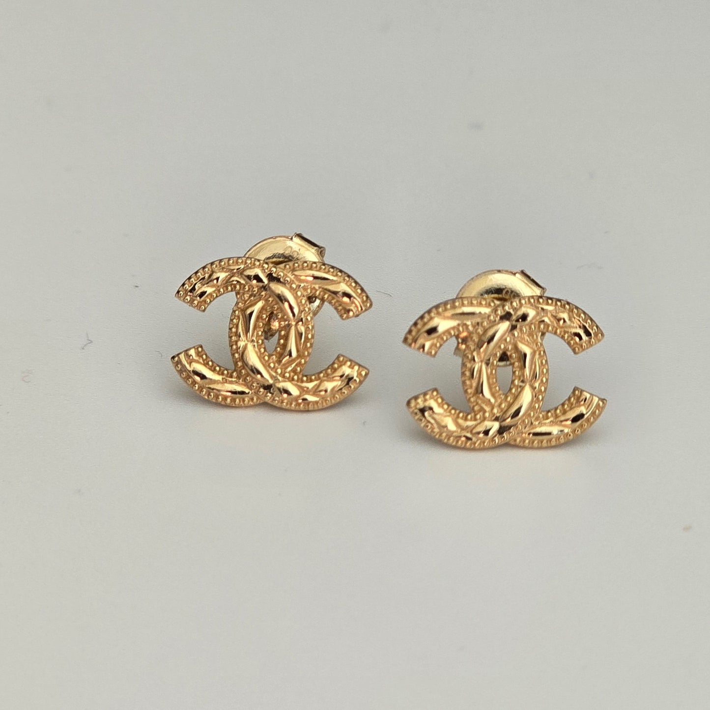 A pair of gold-tone interlocking CC stud earrings with a textured finish, displayed on a light gray background.
