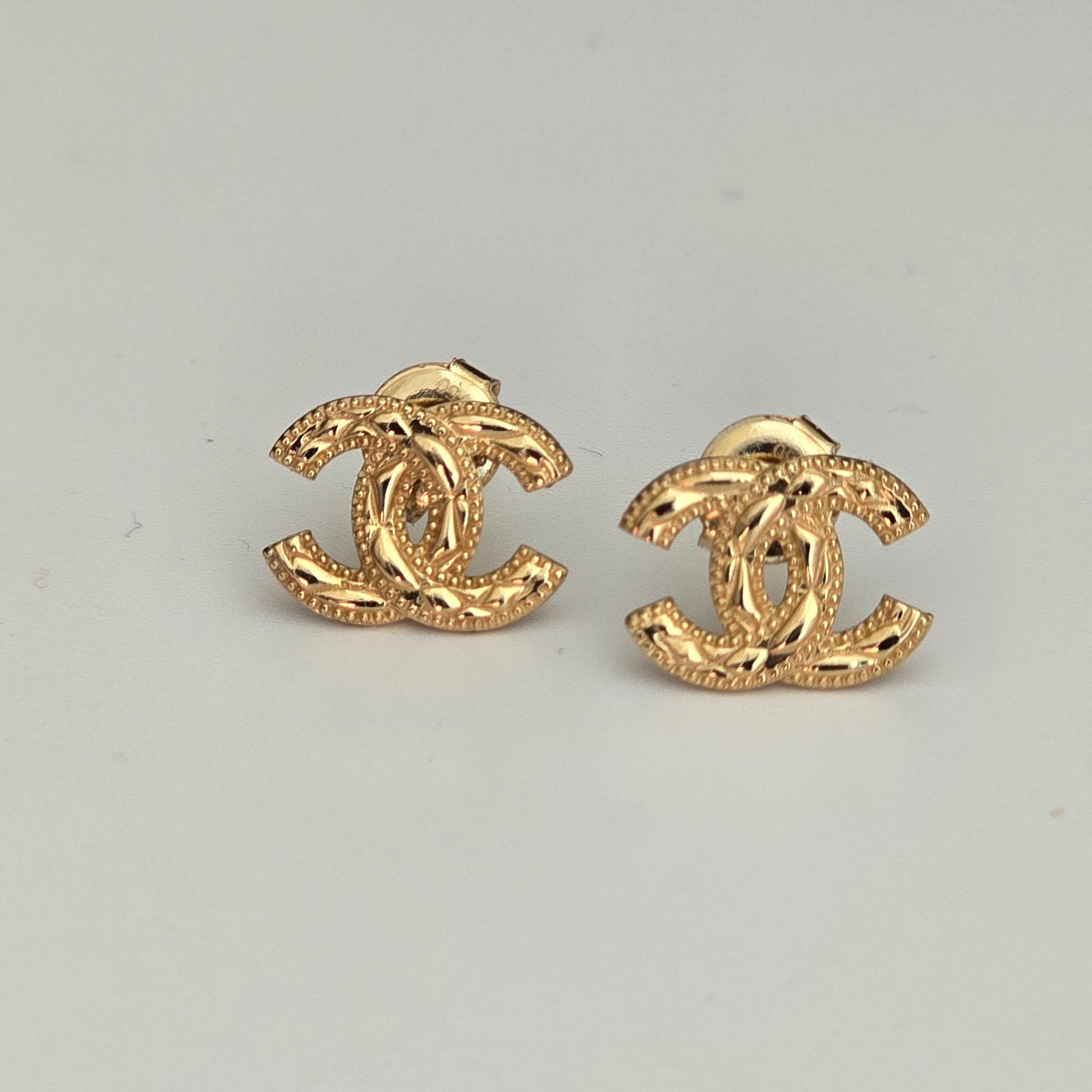 A pair of gold-tone interlocking CC stud earrings with a textured finish, displayed on a light gray background.