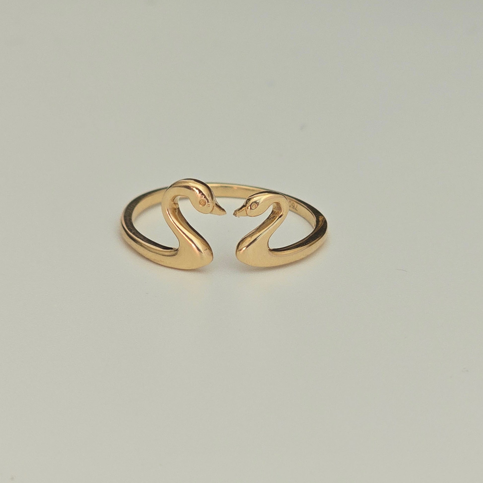 A 18k gold swan ring with two swan heads forming an open band, photographed on a neutral light gray background.