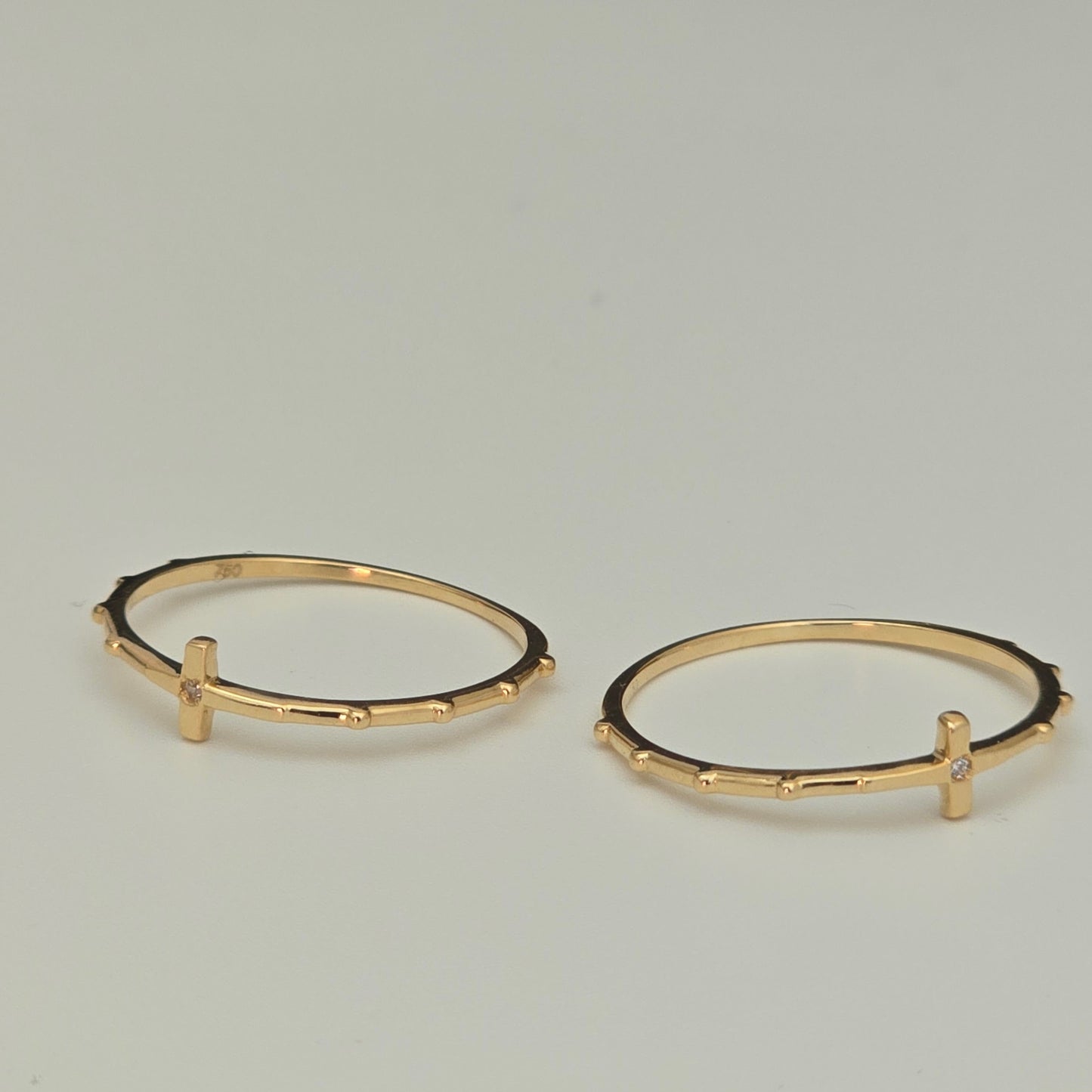 A pair of gold rings with a textured, beaded outer edge and a small vertical bar detail on the front, displayed on a neutral light gray background.