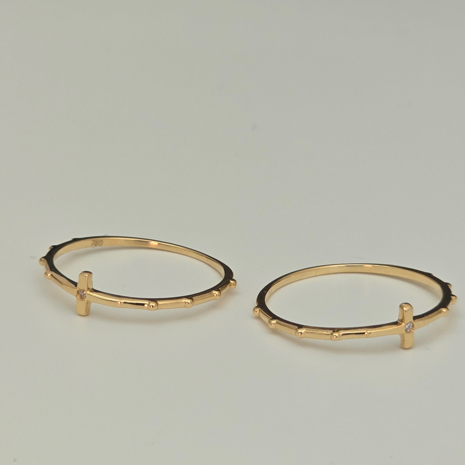 A pair of gold rings with a textured, beaded outer edge and a small vertical bar detail on the front, displayed on a neutral light gray background.