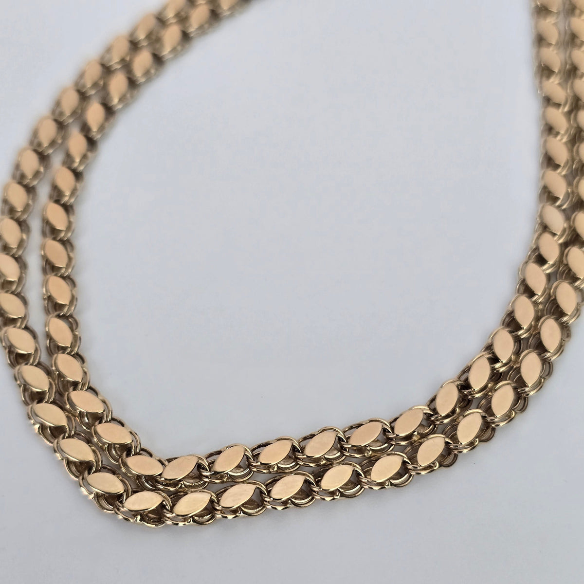 A close-up of a gold-toned multi-strand chain necklace with a textured, polished finish, laid out on a plain light gray background.