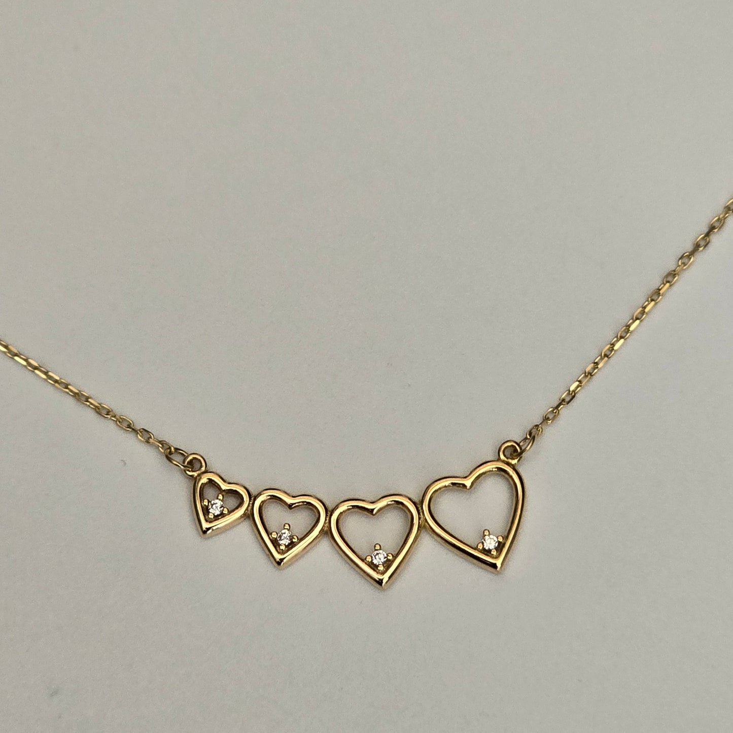 A gold chain necklace featuring three open heart pendants, each adorned with a small clear crystal, displayed against a neutral light gray background.