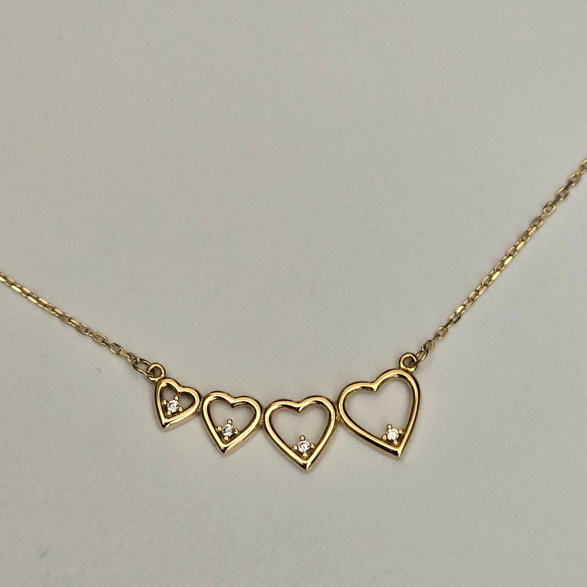 A gold chain necklace featuring three open heart pendants, each adorned with a small clear crystal, displayed against a neutral light gray background.