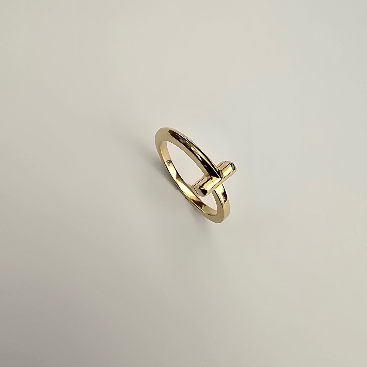 A gold ring with a minimalist, open circular band and a small protruding bar detail, shown on a plain light gray background.
