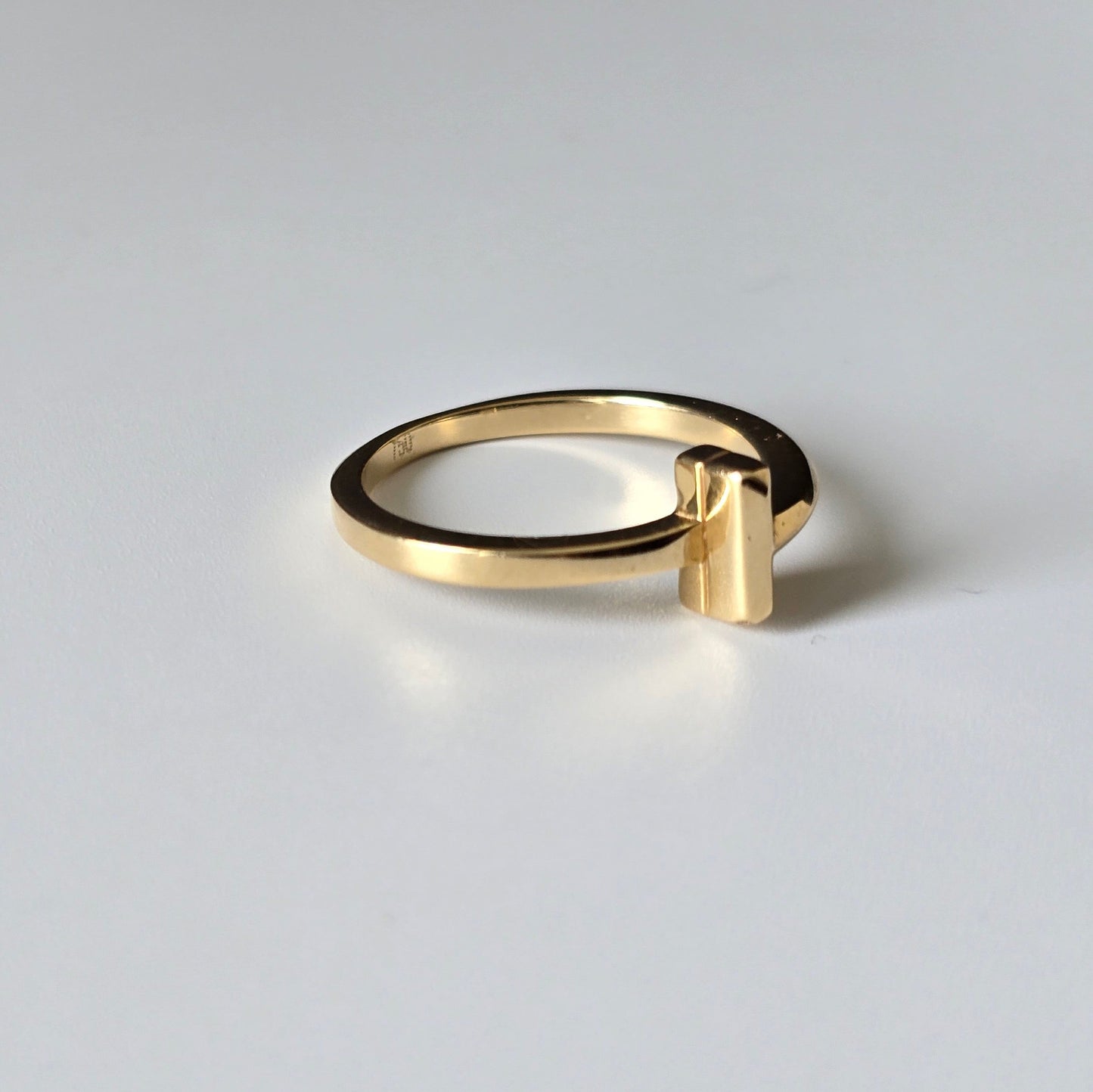 A minimalist gold ring with a wide band and a small rectangular bar extending from the front, photographed on a clean light gray background.
