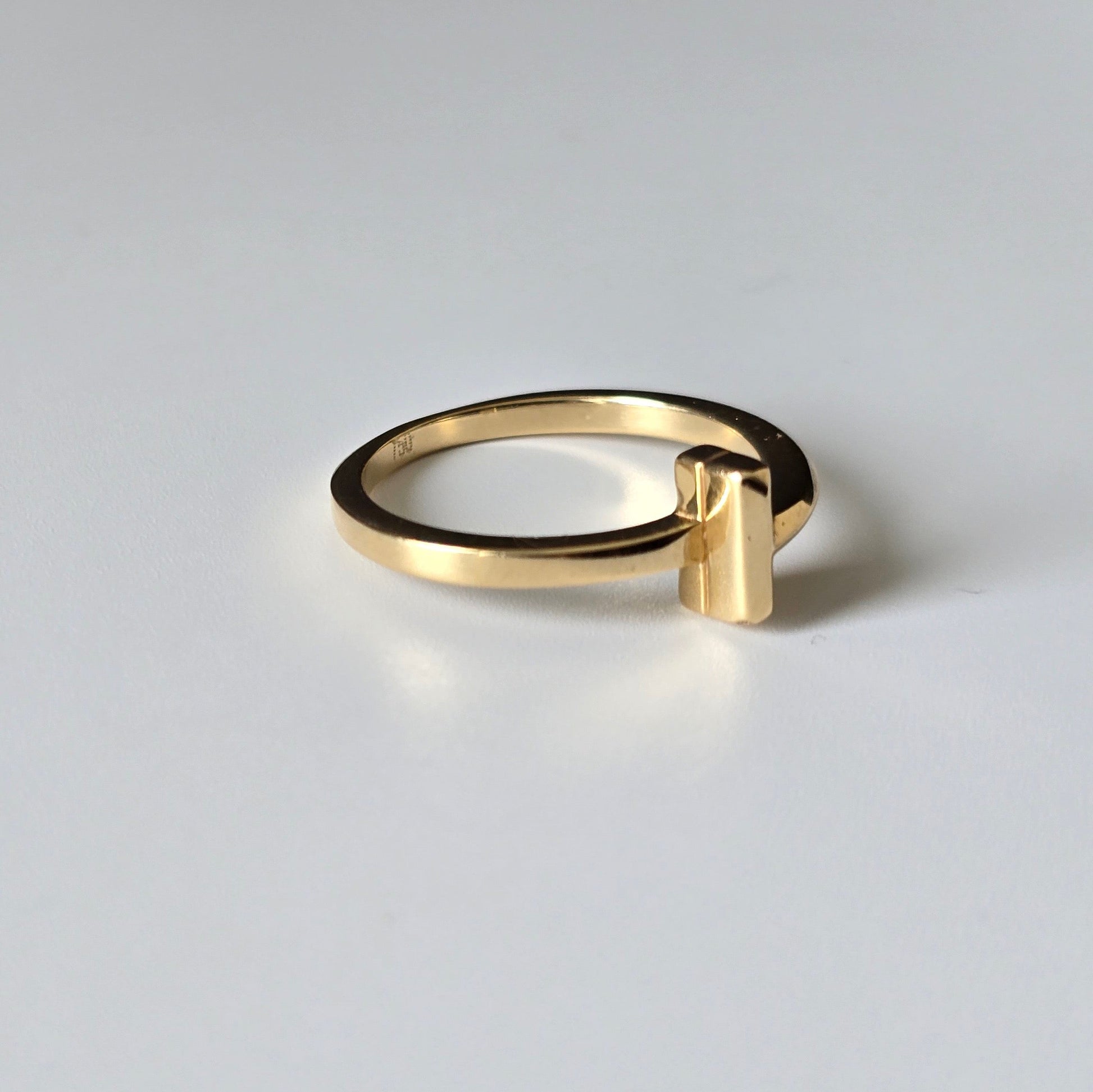 A minimalist gold ring with a wide band and a small rectangular bar extending from the front, photographed on a clean light gray background.