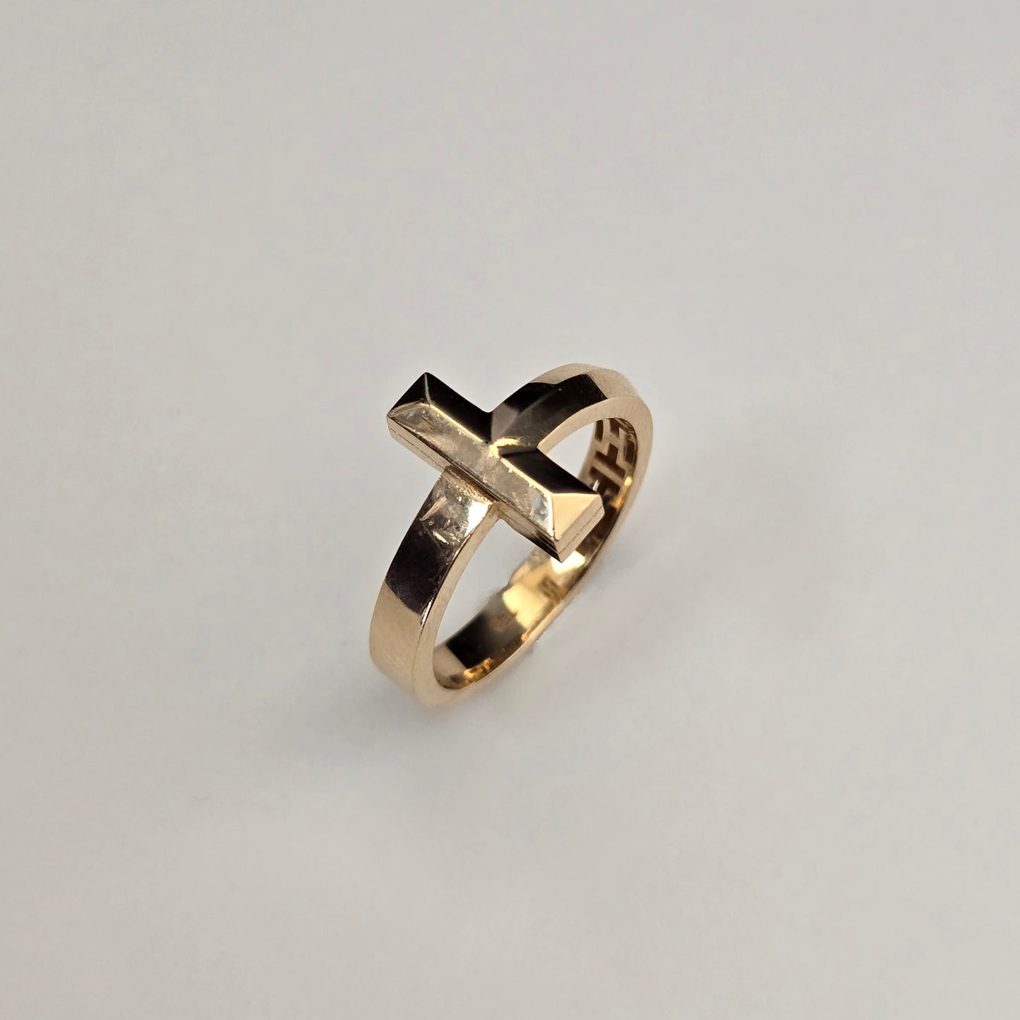 A gold ring with a geometric, faceted cross-like top and a small clear inset stone, shown on a plain light gray background.