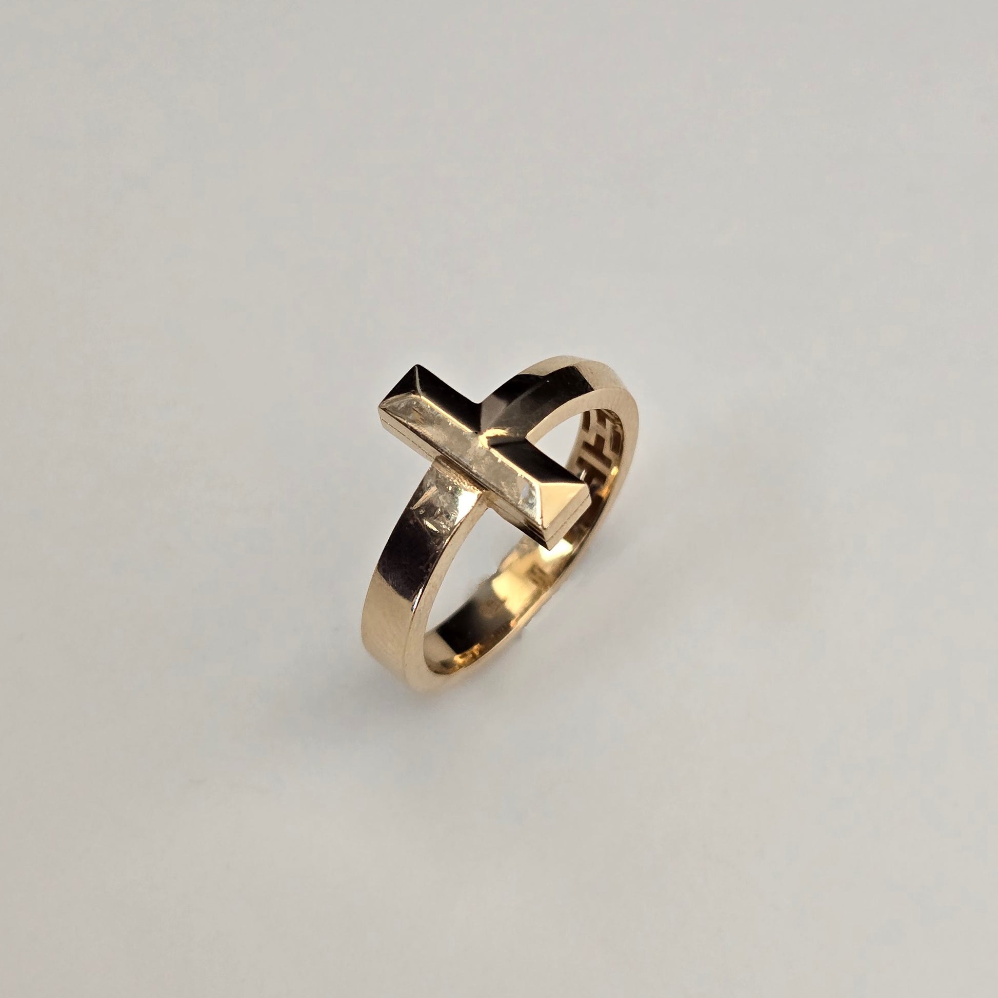 A gold ring with a geometric, faceted cross-like top and a small clear inset stone, shown on a plain light gray background.