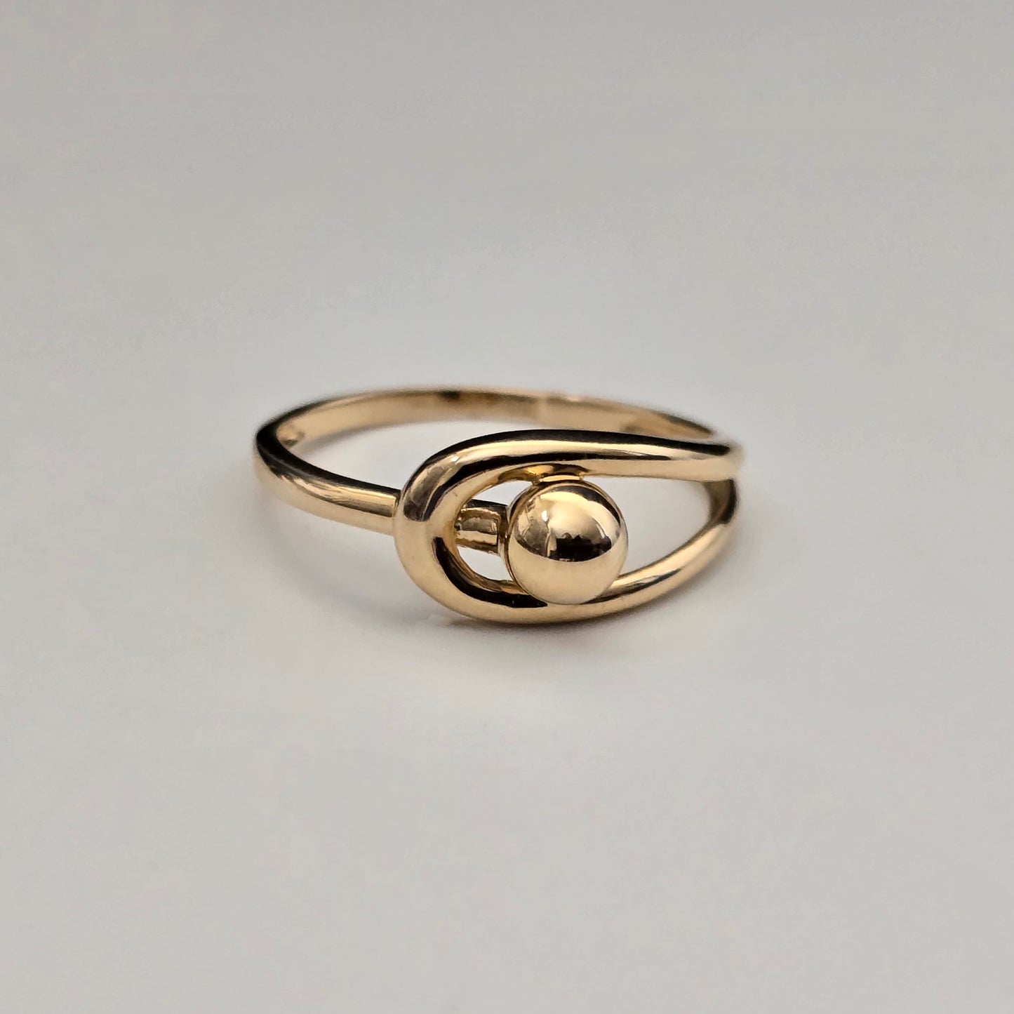 A gold-colored ring with a minimalist, abstract design featuring a polished oval loop and a central spherical bead, set against a plain light gray background.