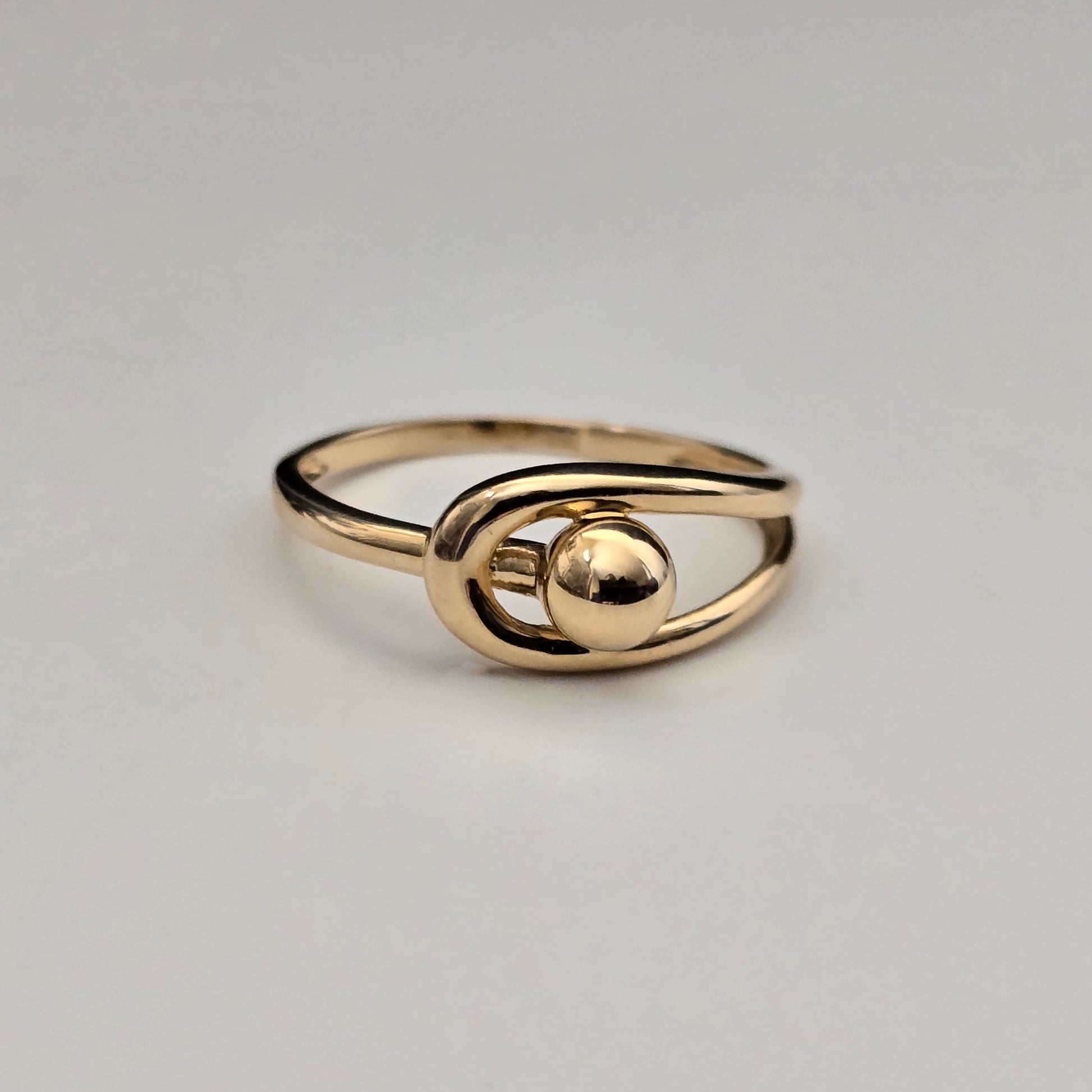 A gold-colored ring with a minimalist, abstract design featuring a polished oval loop and a central spherical bead, set against a plain light gray background.