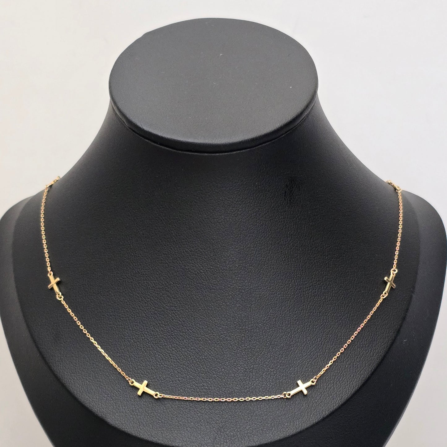 A delicate gold chain necklace with small cross charms spaced along the front, displayed on a matte black jewelry bust against a light gray background.