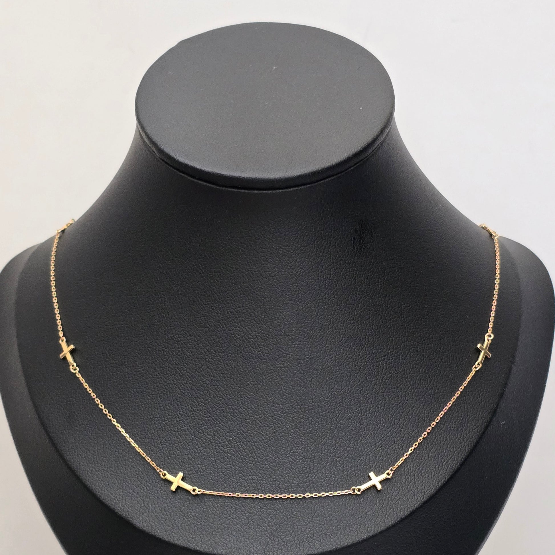 A delicate gold chain necklace with small cross charms spaced along the front, displayed on a matte black jewelry bust against a light gray background.