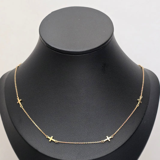 A delicate gold chain necklace with small cross charms spaced along the front, displayed on a matte black jewelry bust against a light gray background.