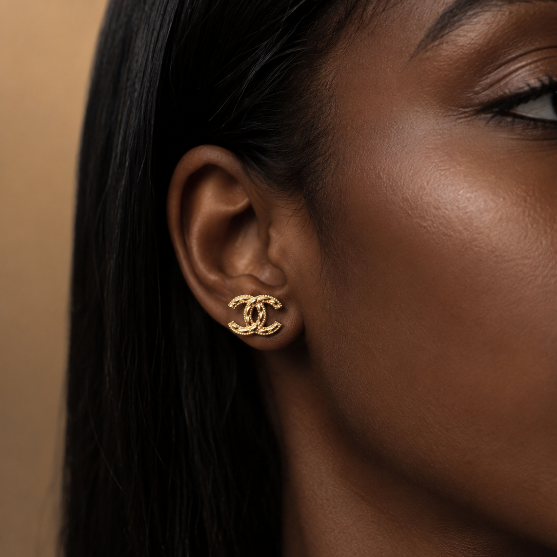 A close-up side profile of a woman with dark hair wearing a small gold double-C stud earring, showing the Chanel logo.